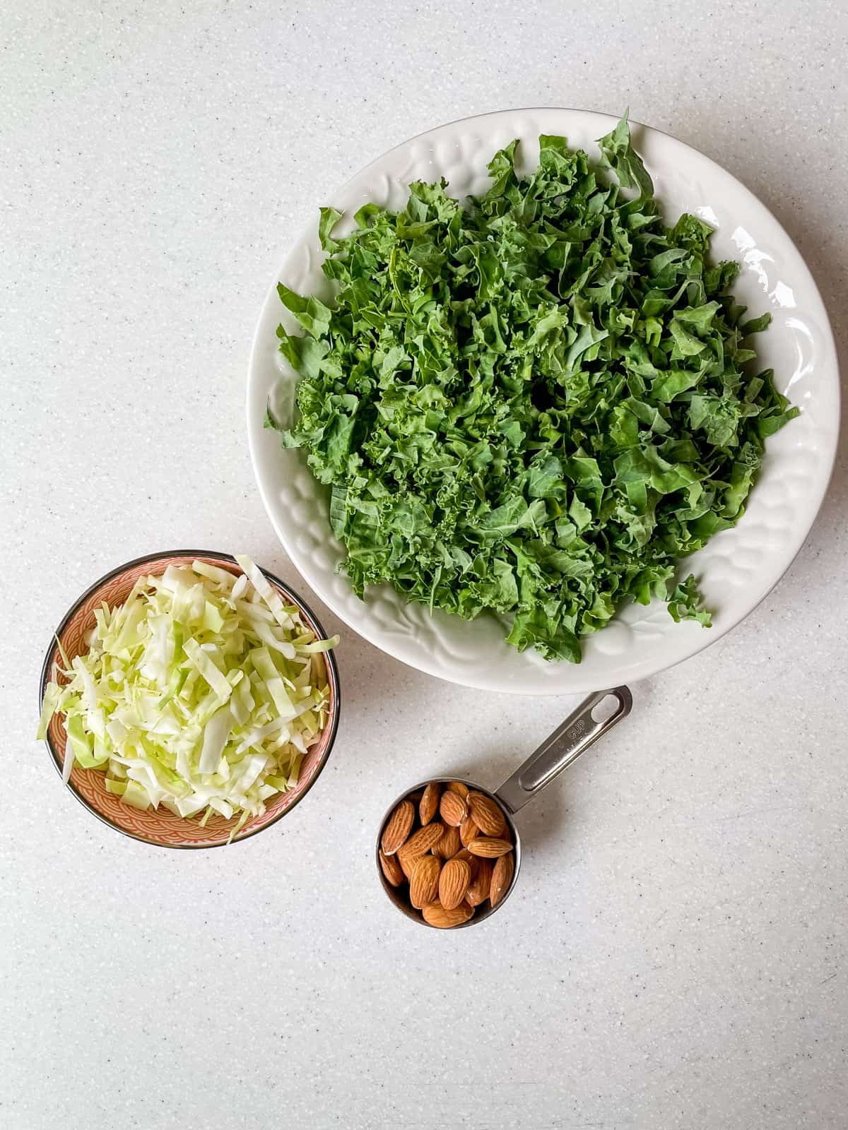 finely chopped kale in bowl, next to shredded green cabbage and almonds.