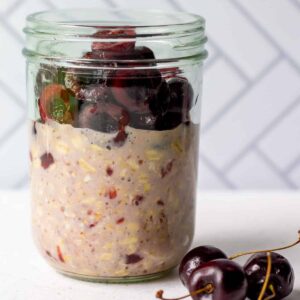 mason jar with cherry flavored overnight oats and fresh pitted halved cherries on top, with three cherries on the counter beside the jar