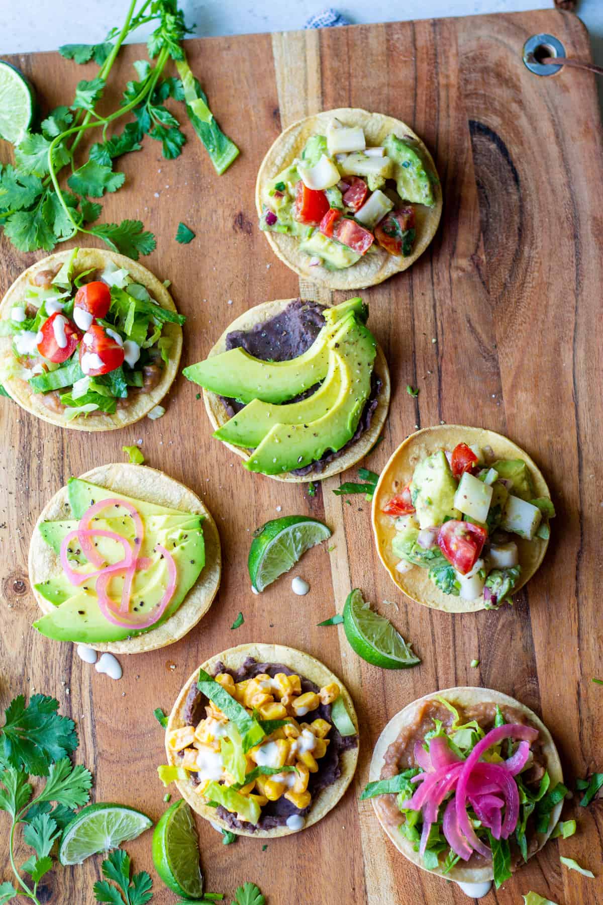 seven different mini tostadas with different plant-based toppings like street corn, vegan ceviche, beans, avocado, pickled onion and lettuce on a wooden board