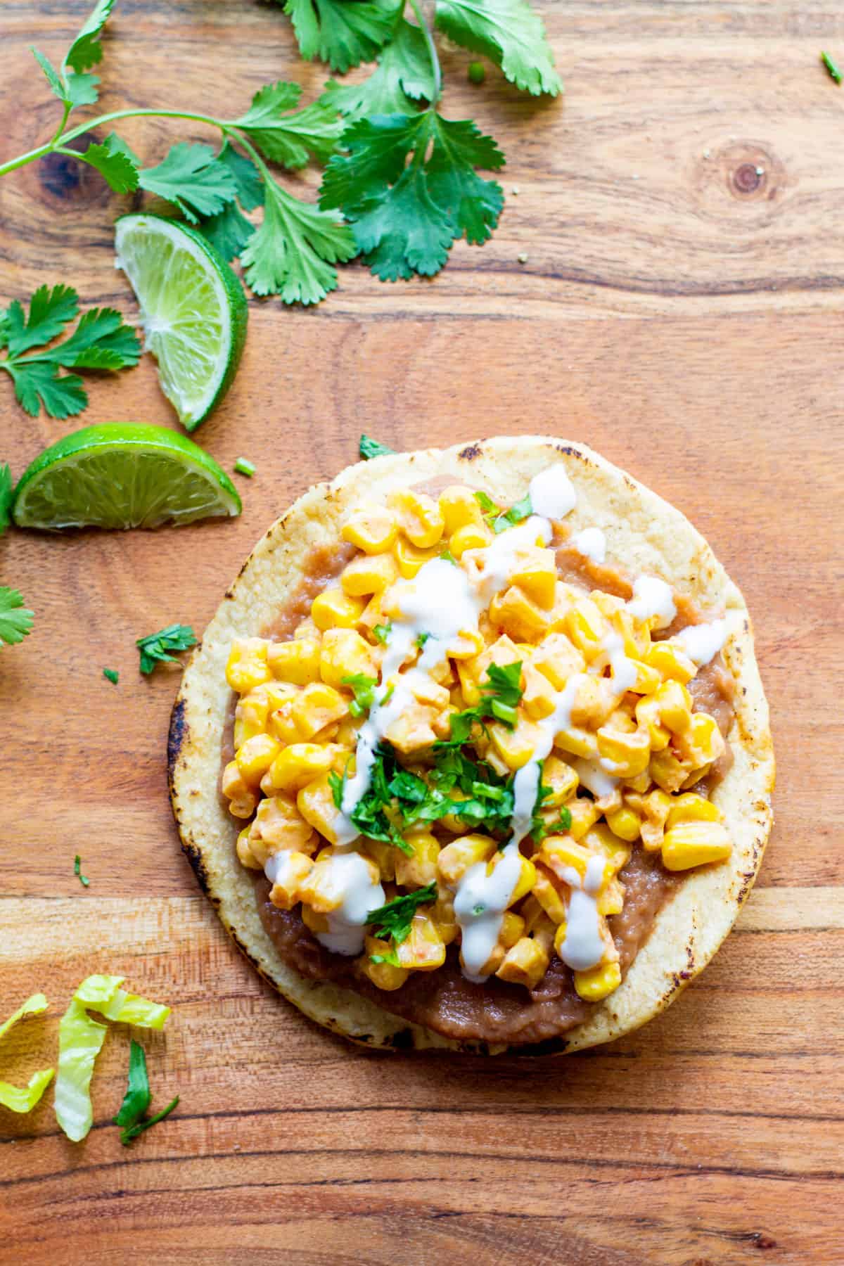 tostada with refried beans, dairy-free mexican street corn esquites, sour cream and cilantro on a wooden board with a lime in the background