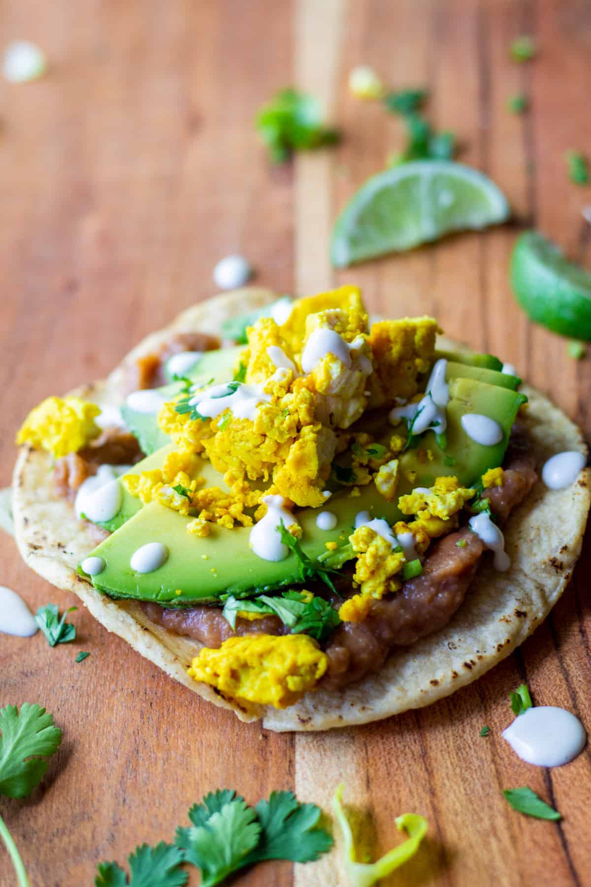 tostada with refried beans, sliced avocado, tofu scramble, crema and cilantro on a wooden board