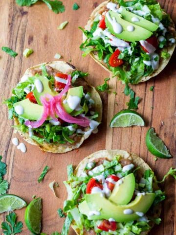Three tostadas on top of a wooden cutting board with refried beans, lettuce, avocado, red onion, and tomato with lime wedges on the side.
