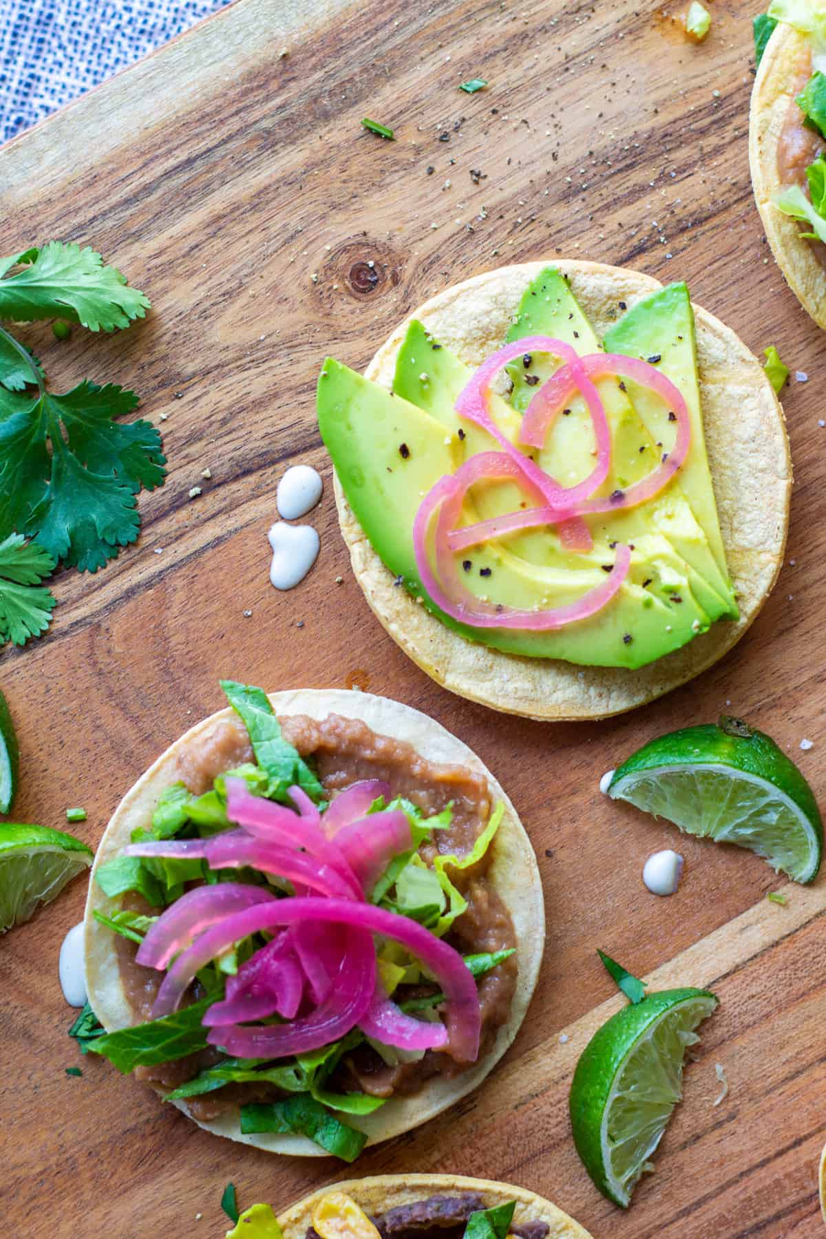 mini vegan tostada with avocado, pickled onion, beans, lettuce on wooden board with cut limes and cilantro on the side
