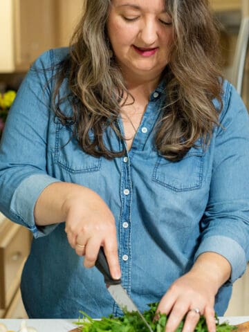 woman in jean shirt chopping cilantro on wooden cutting board, with garlic and lemon nearby