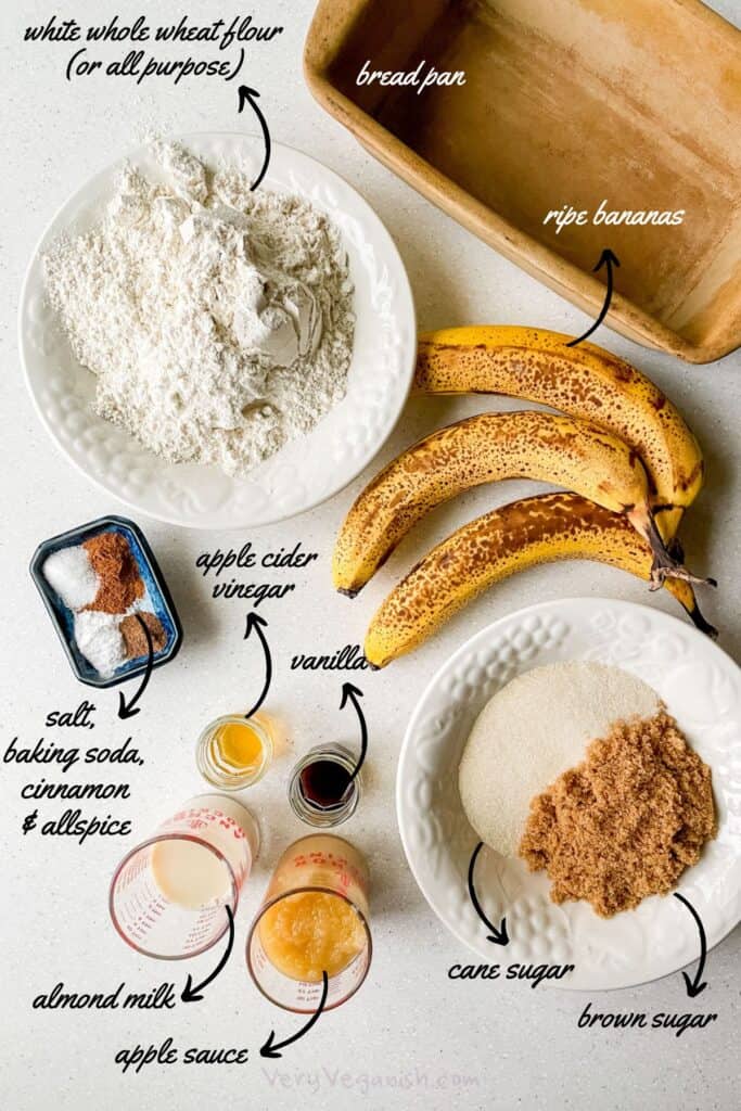 Ingredients for vegan banana bread with applesauce measured out on counter: white whole wheat flour, ripe bananas, cane sugar, brown sugar, apple sauce, almond milk, vanilla, apple cider vinegar, salt, baking soda, cinnamon and allspice