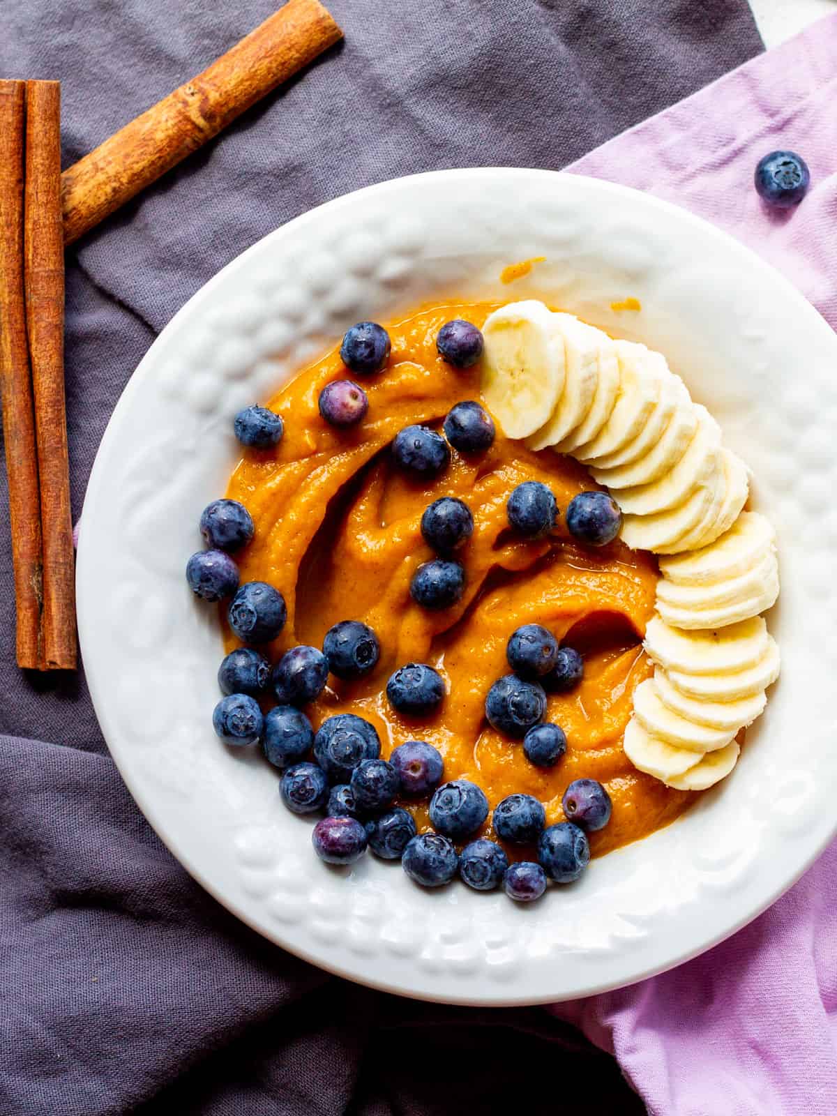 white bowl of pureed vegan sweet potato pudding with sliced bananas and fresh blueberries on top with cinnamon sticks on table next to bowl