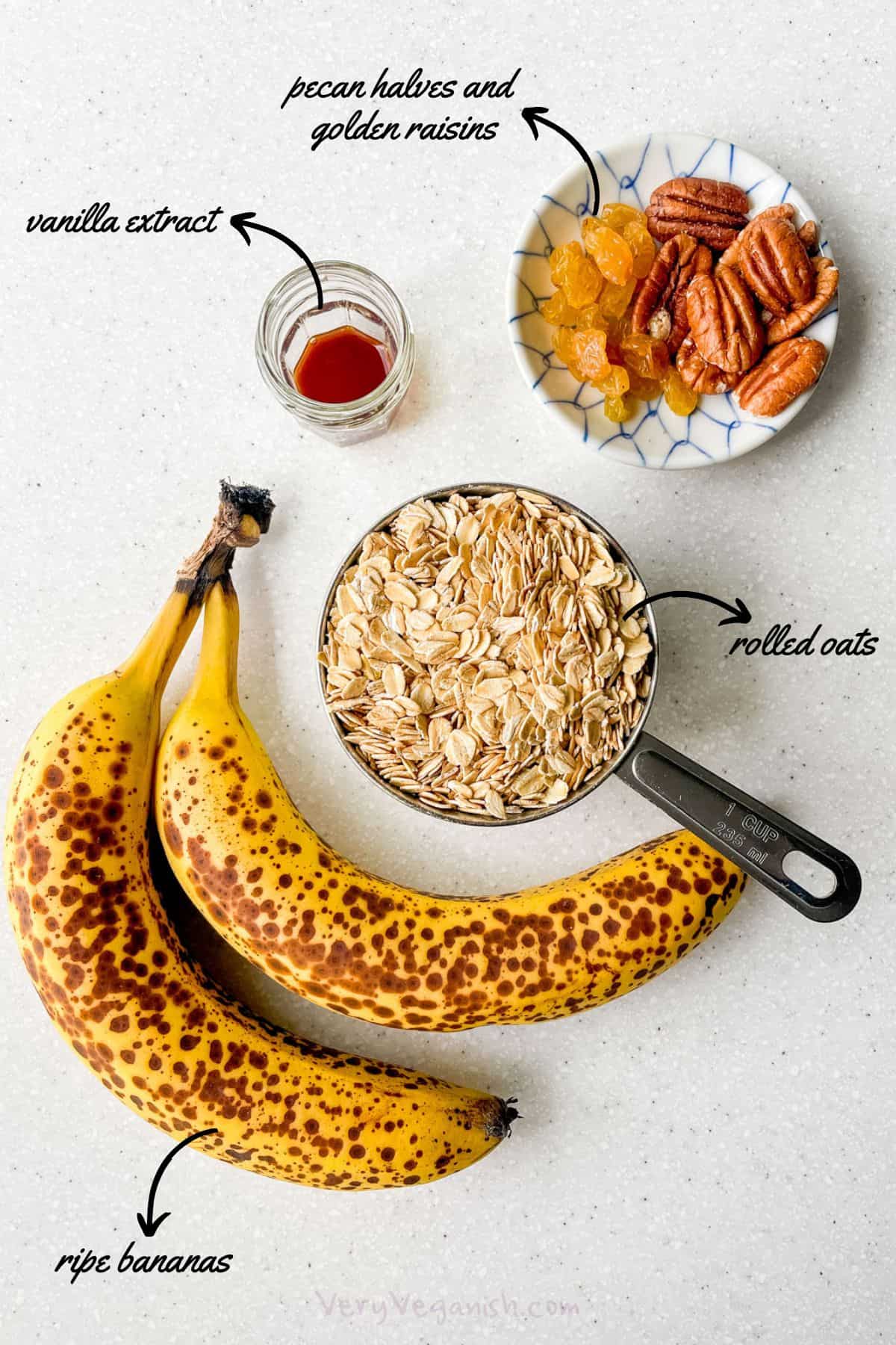 Ingredients for simple banana oatmeal cookies with pecans and golden raisins on a counter: ripe bananas, rolled oats, vanilla extract, pecan halves and golden raisins.