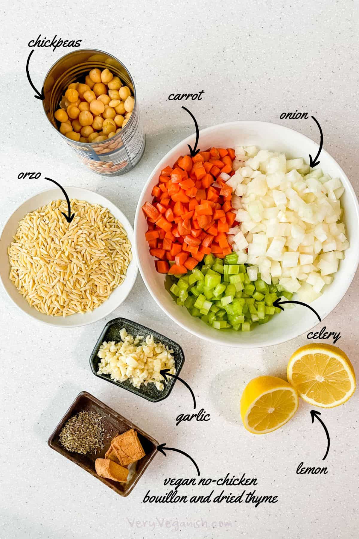 Ingredients for Chickpea Lemon Orzo Soup. Canned chickpeas, a white bowl with orzo pasta inside, a bigger white bowl with thirds full of diced carrots, chopped onion, and chopped celery to the left. A small dish in the middle of the screen contains some minced garlic, and there are two halves of lemon beside the dish to the left. There is another small dish with two cubes of vegan no-chicken bouillon and a small pile of dried thyme.
