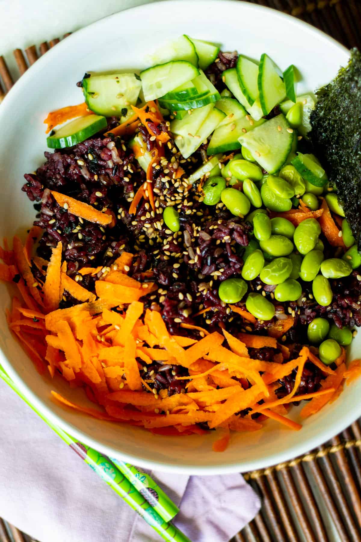 white bowl of dark purple sushi rice with cucumber, edamame, carrot, seaweed snacks and sesame seeds