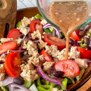 pouring Greek salad dressing from a glass measuring cup onto a vegan greek salad in a big wooden bowl