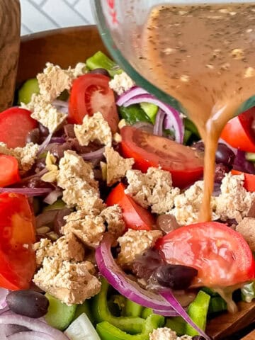 pouring Greek salad dressing from a glass measuring cup onto a vegan greek salad in a big wooden bowl