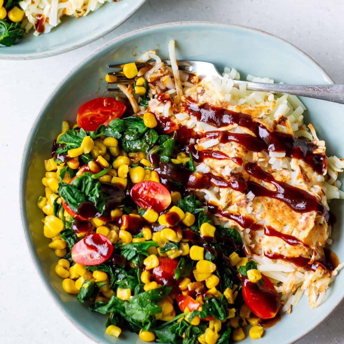 light blue bowl with golden hash browns and cooked spinach, corn and tomato on the side with BBQ sauce drizzled on top and fork in the bowl