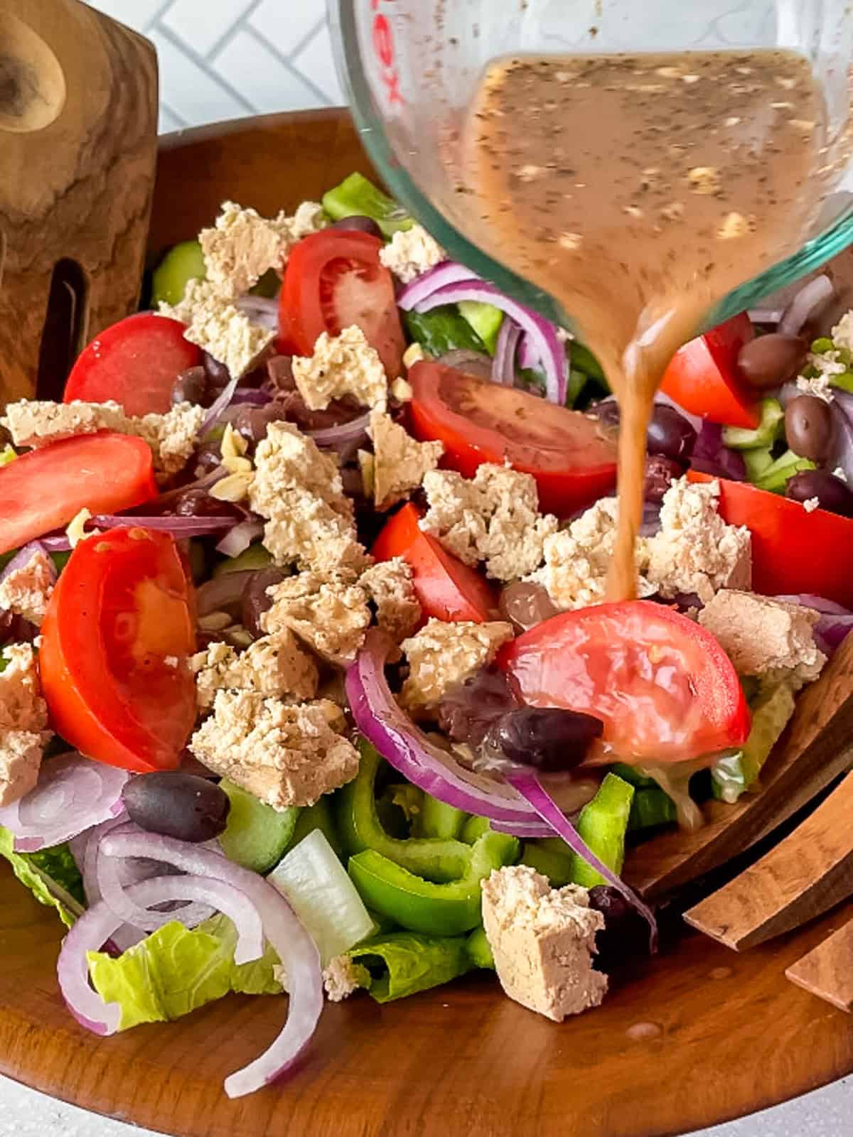 pouring Greek salad dressing from a glass measuring cup onto a vegan greek salad in a big wooden bowl
