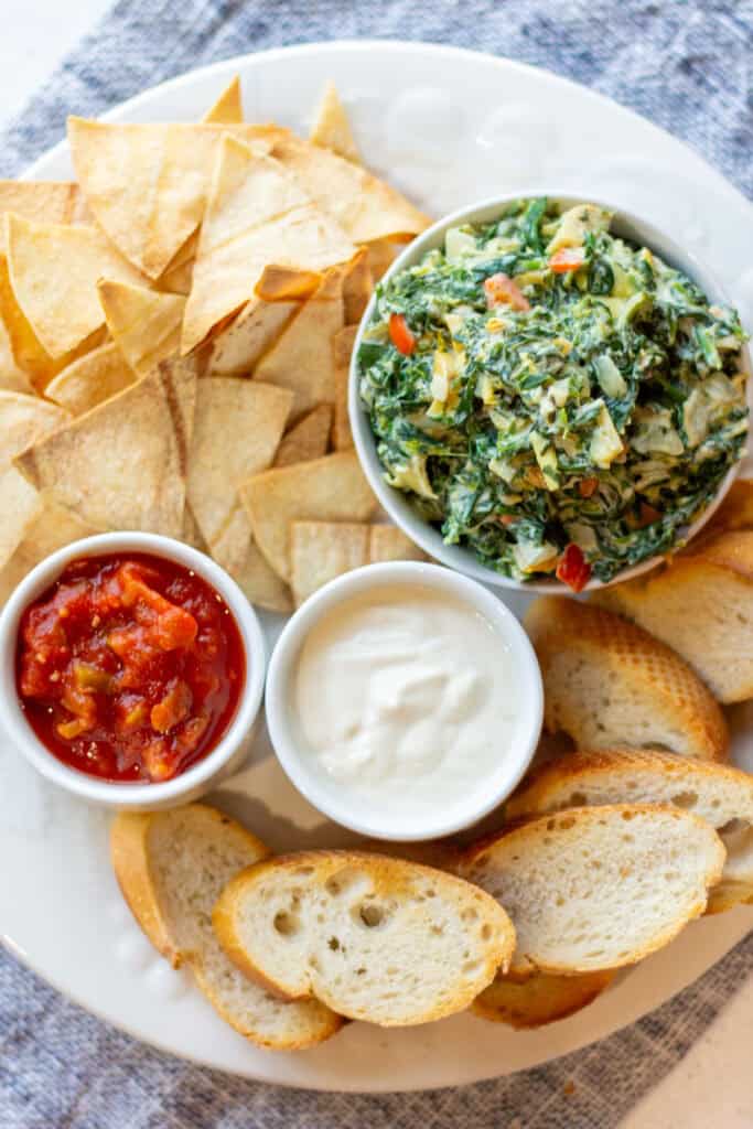 white plate on a blue placemat with tortilla chips, toasted french bread, vegan spinach artichoke dip, dairy-free sour cream and tomato salsa
