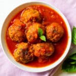 six eggplant meatballs with marinara sauce in a white bowl over a purple napkin with fresh basil garnish and fresh basil leaves on table