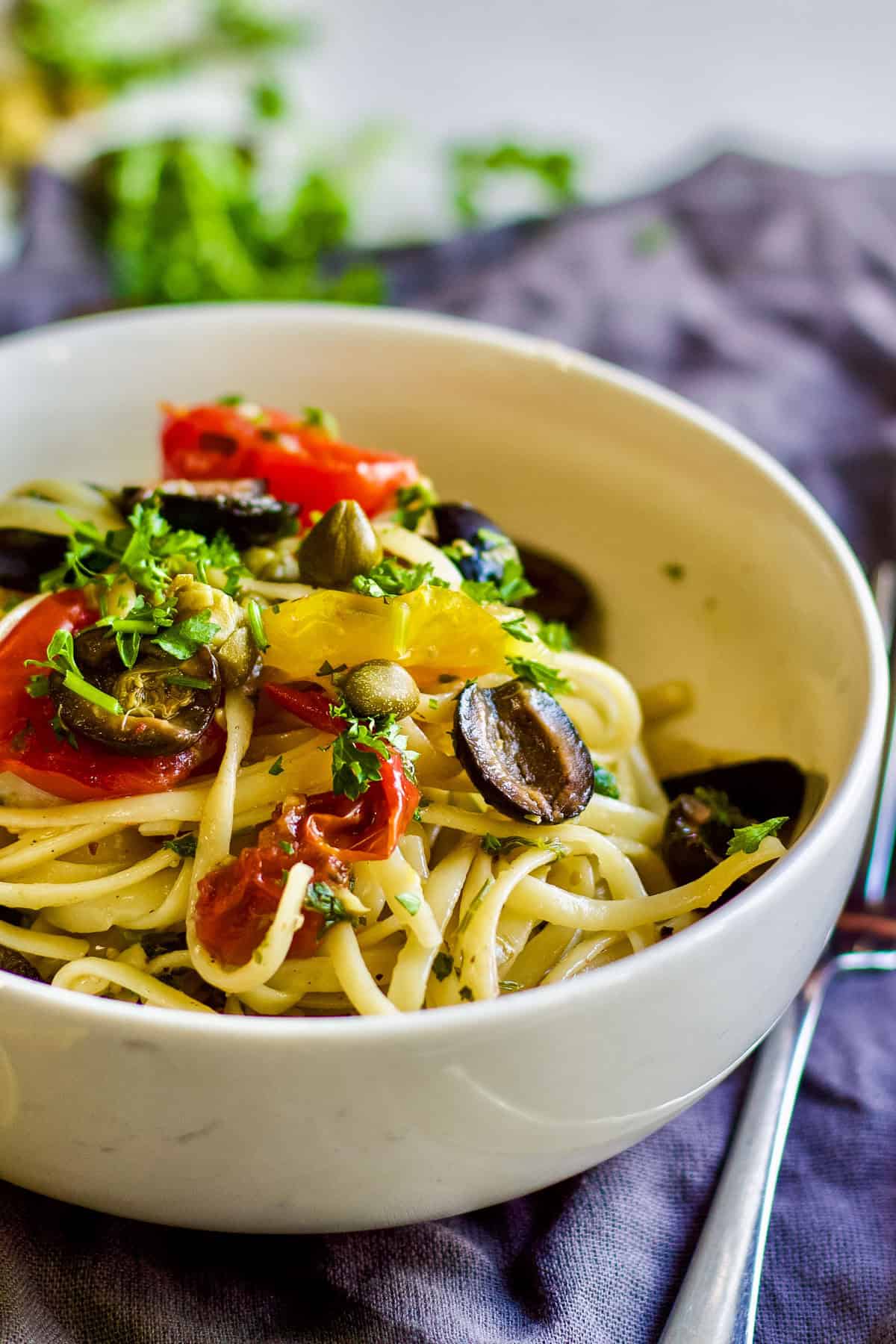 white bowl of pasta puttanesca with sliced black olives, capers, tomato and parsley on top