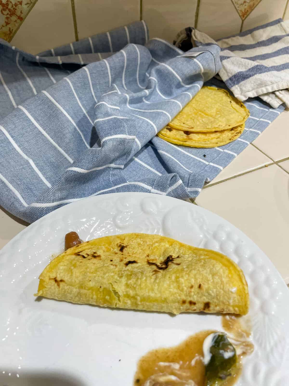 Corn tortilla (heated over open flame) filled with beans and sour cream on white plate with more tortillas in tea towel