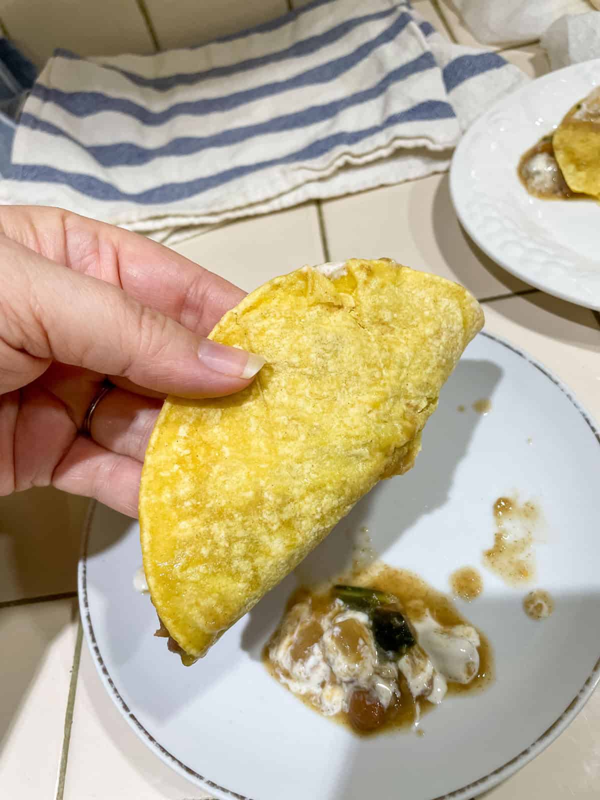 Corn tortilla (heated in non-stick skillet with steam) filled with beans and sour cream on white plate with more tortillas in tea towel