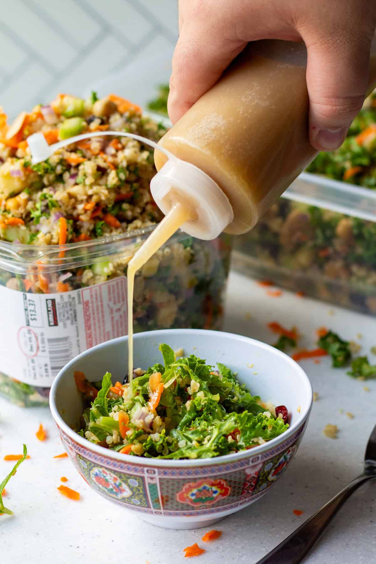 Pouring homemade apple cider dijon vinaigrette dressing over grain and celery copycat Costco salad with a Kirkland plastic container full of salad in background