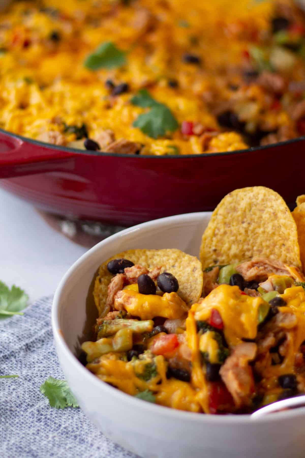 White bowl of cheesy soy curl, black bean, corn and broccoli taco mixture with the skillet in the background and cilantro for garnish