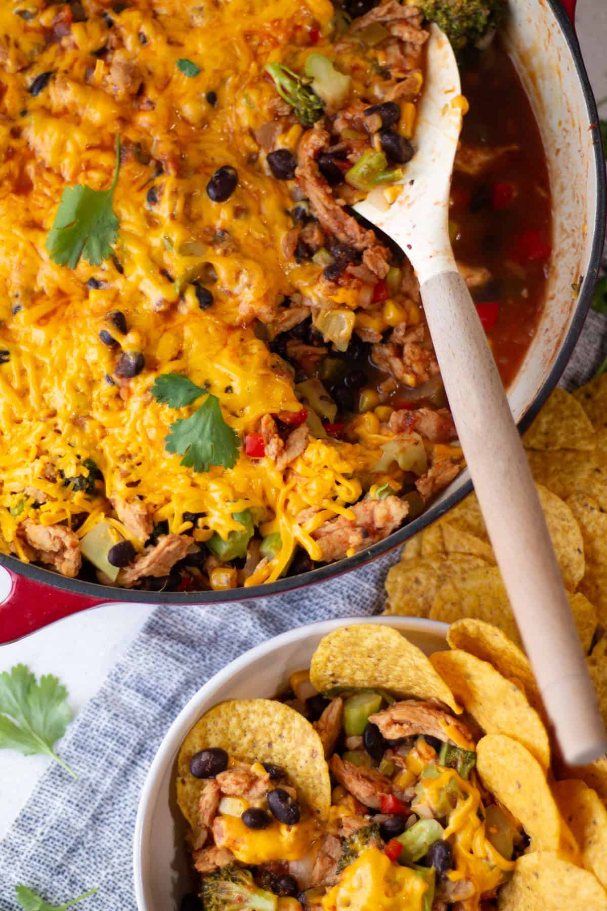 Scooping the cheesy soy curl, black bean, corn and broccoli taco mixture from a wide skillet with tortilla chips on the table near a bowl