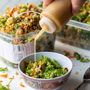 Pouring homemade apple cider dijon vinaigrette dressing over grain and celery copycat Costco salad with a Kirkland plastic container full of salad in background