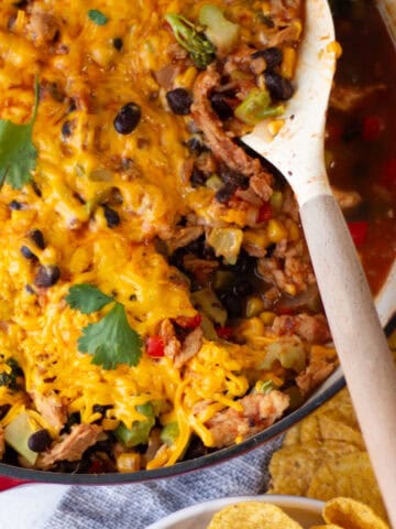 Scooping the cheesy soy curl, black bean, corn and broccoli taco mixture from a wide skillet with tortilla chips on the table near a bowl