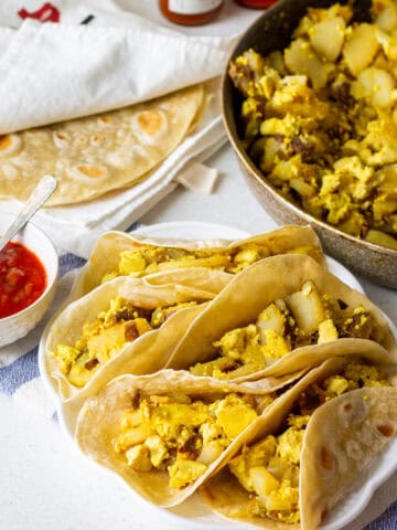 Cooked Tortilla Land flour tortillas keeping warm inside a tea towel with vegan potato and egg tacos on a plate and in the skillet with salsa as well