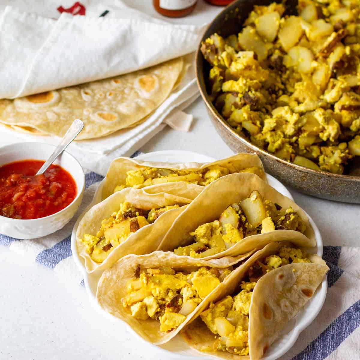 Cooked Tortilla Land flour tortillas keeping warm inside a tea towel with vegan potato and egg tacos on a plate and in the skillet with salsa as well