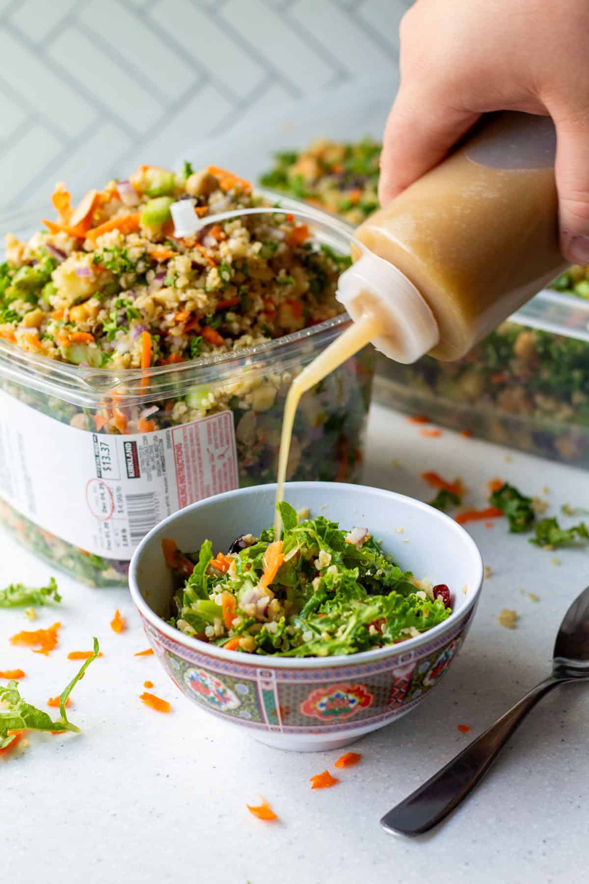 Pouring homemade apple cider vinaigrette dressing over grain and celery copycat Costco salad with a Kirkland plastic container full of salad in background