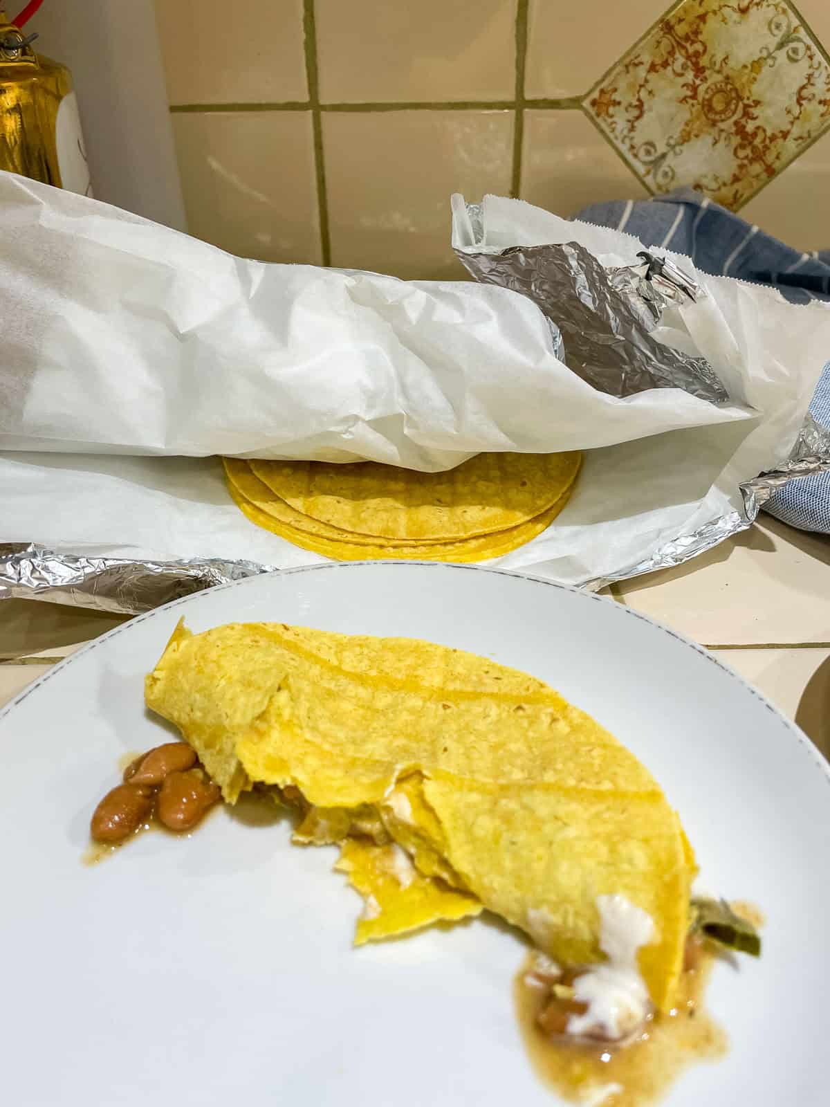 Corn tortilla (heated in oven) filled with beans and sour cream on white plate with more tortillas in tea towel