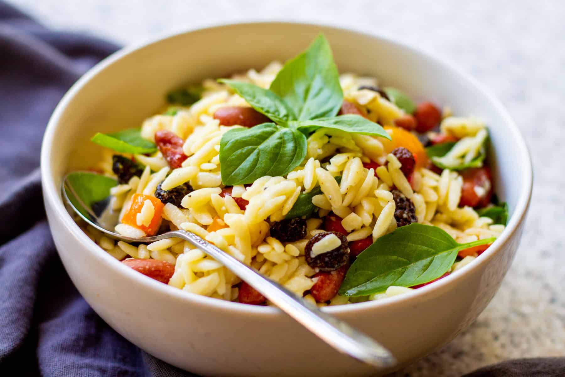 white bowl filled with orzo basil kidney bean vegetable pasta salad with a sprig of fresh basil and a spoon resting on top