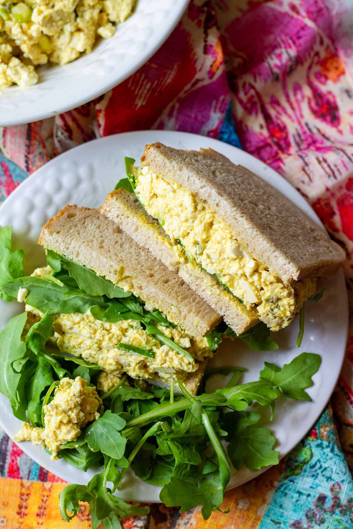 Prepared tofu egg salad sandwich with arugula on white plate and colorful napkin underneath