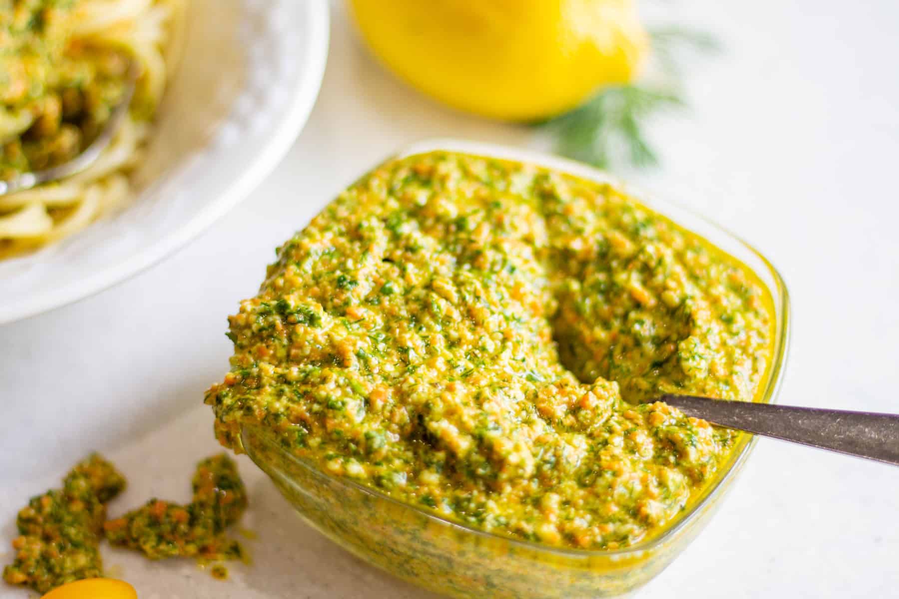 bowl of dill parsley pesto with a spoon nestled inside. Bowl of pasta, a lemon, some fresh dill and some tomatoes nearby.