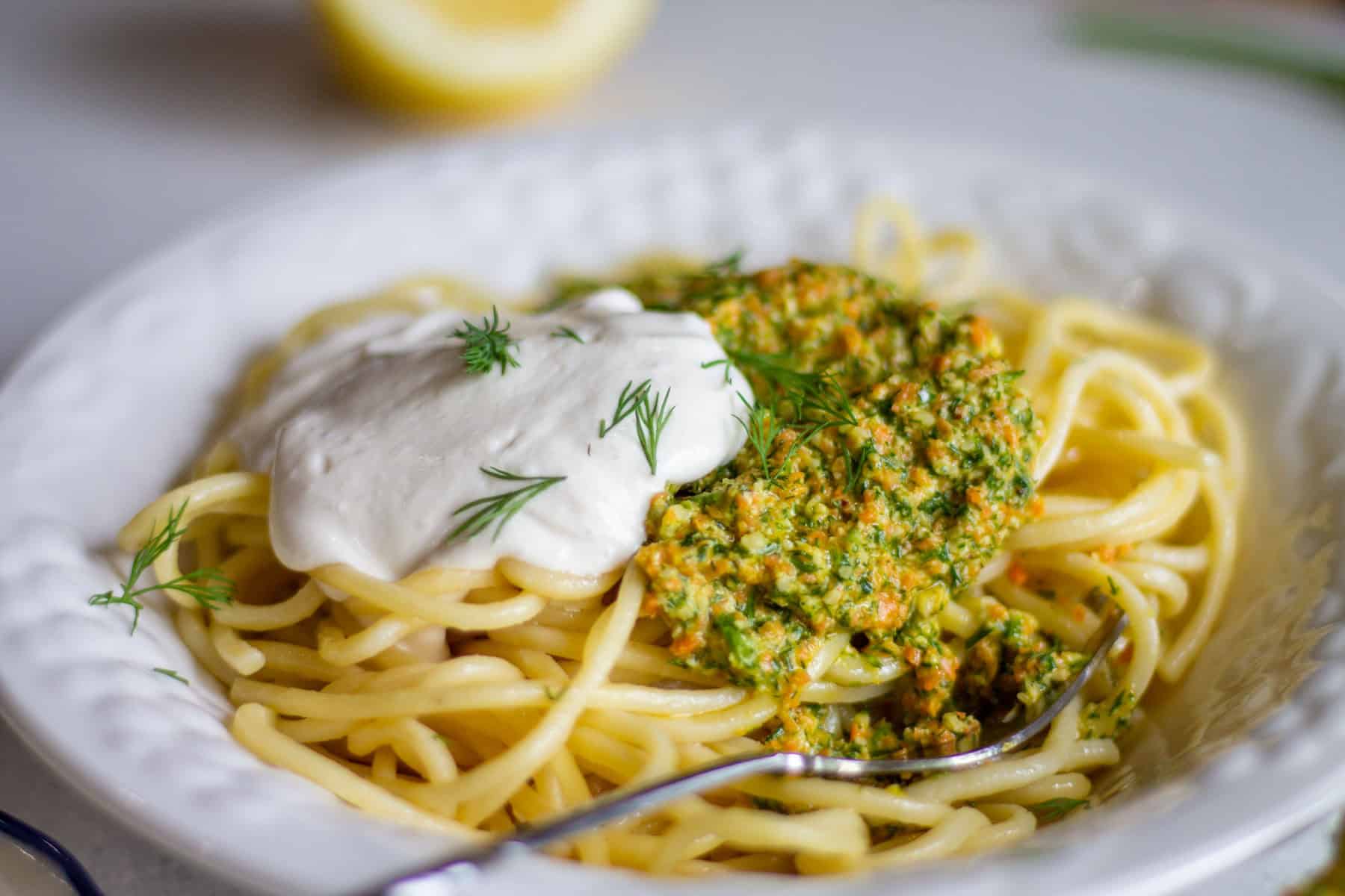Spaghetti with dill parsley pesto and vegan sour cream in a white bowl with a fork on the side