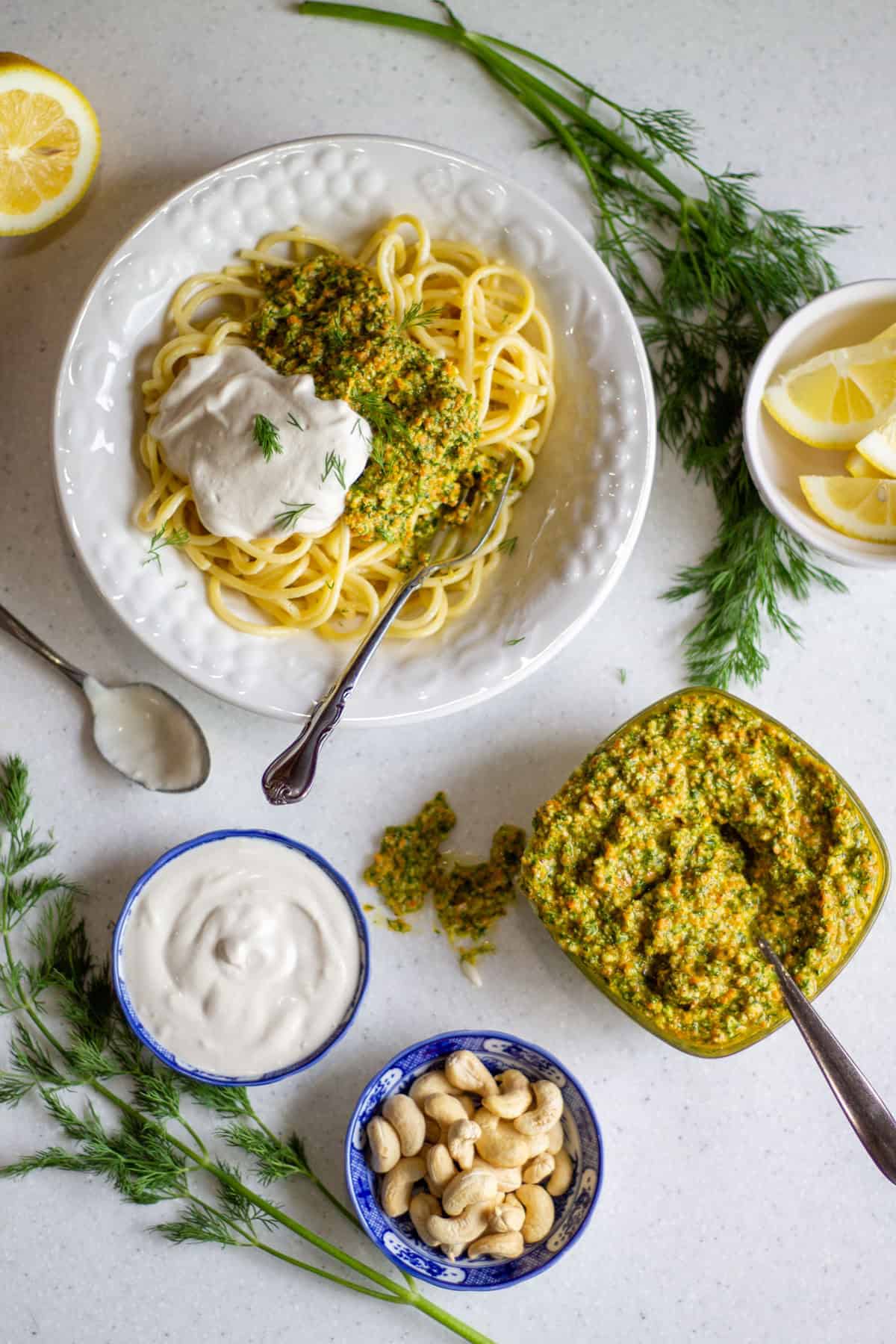 Spaghetti with dill parsley pesto and vegan sour cream in a white bowl with a fork on the side. Also on the counter is a bowl of pesto, bowl of sour cream, bowl of cashews, lemon wedges and fresh dill.