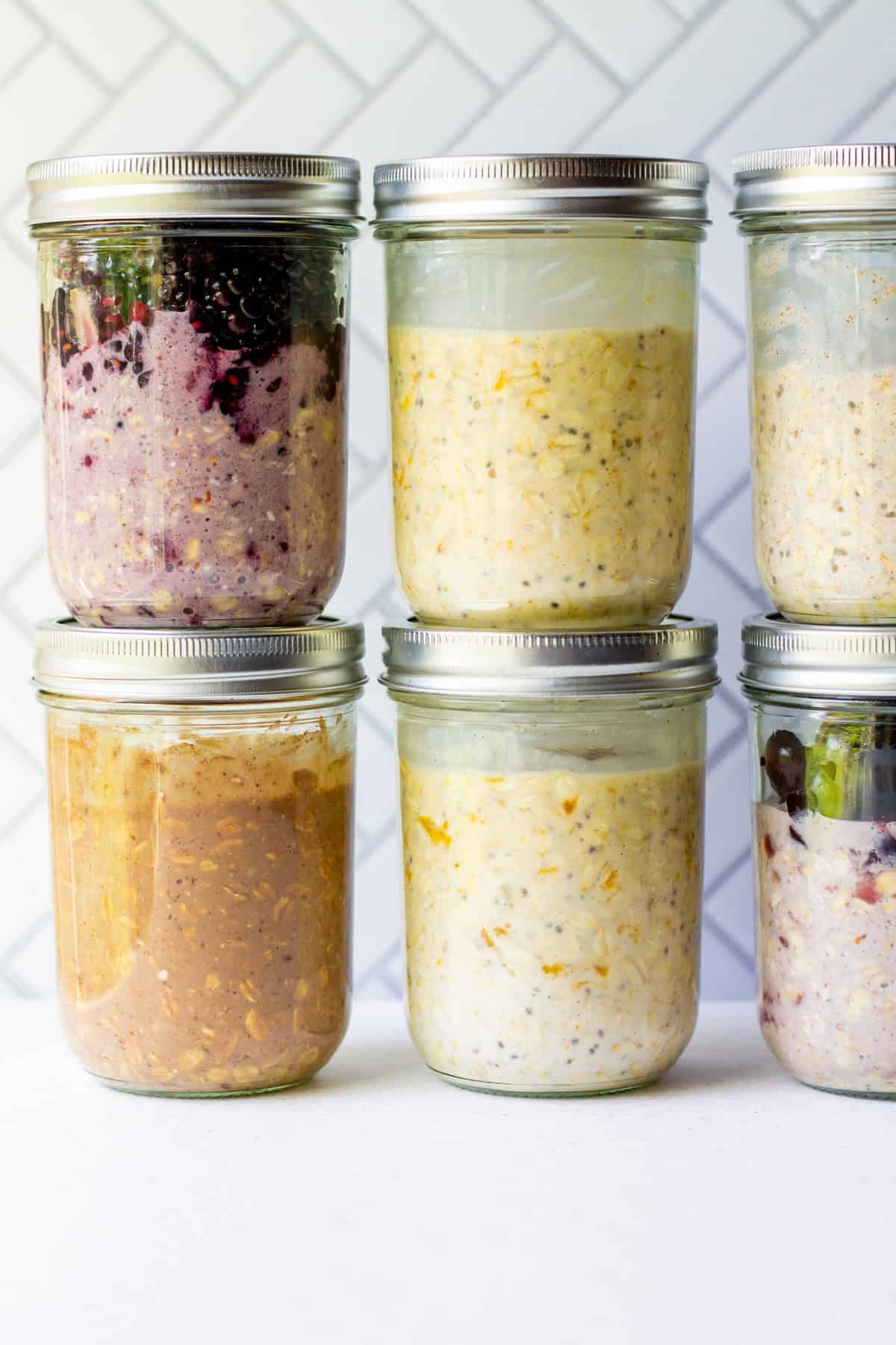 Six jars of vegan overnight oats in different flavors arranged in two rows, including blackberry, double chocolate, apricot, apricot cardamom, vanilla, and cherry, perfect for meal prep.