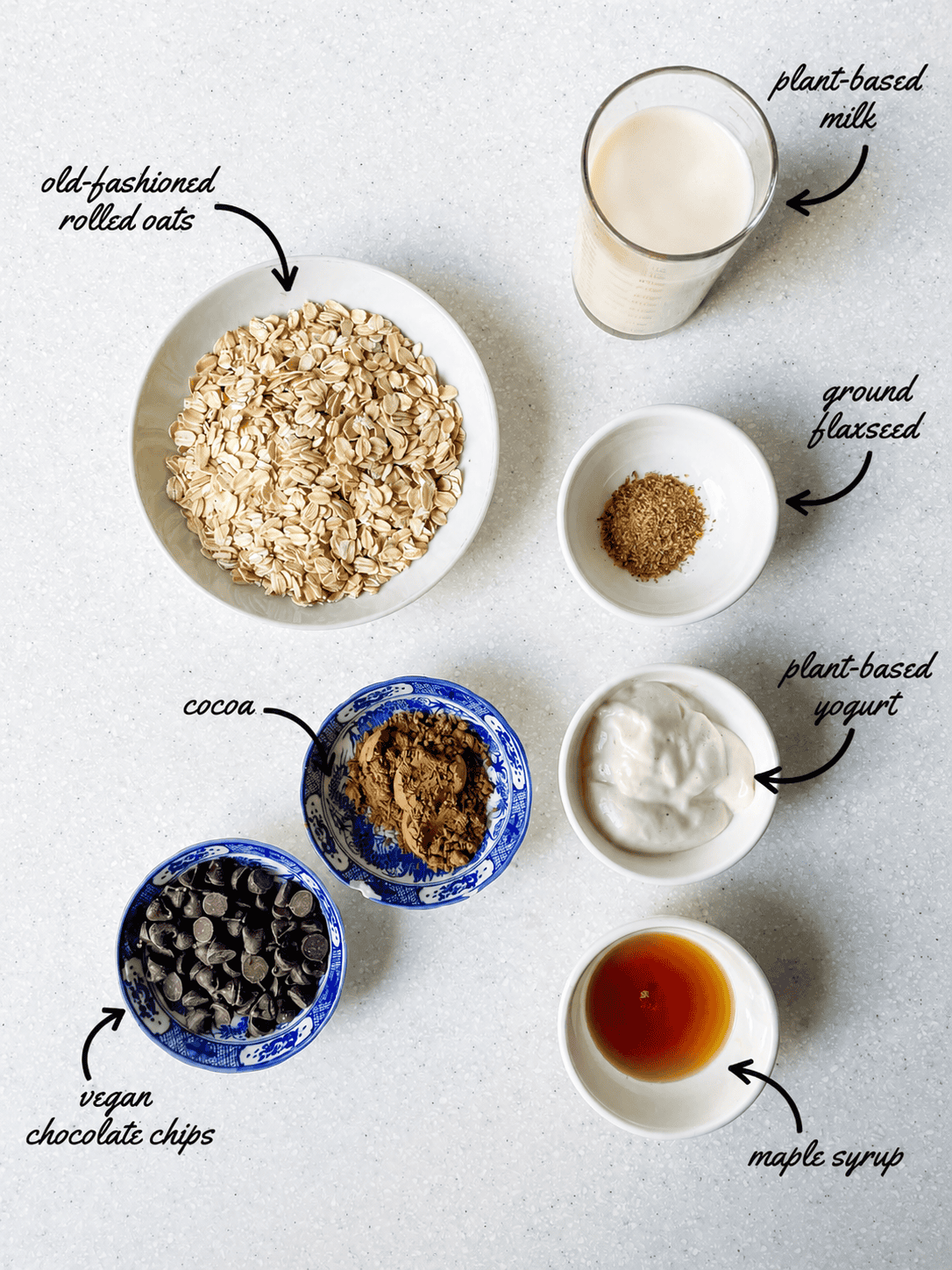 Overhead view of ingredients for vegan double chocolate overnight oats, including rolled oats, plant-based milk, ground flaxseed, plant-based yogurt, cocoa powder, vegan chocolate chips, and maple syrup, labeled with arrows.