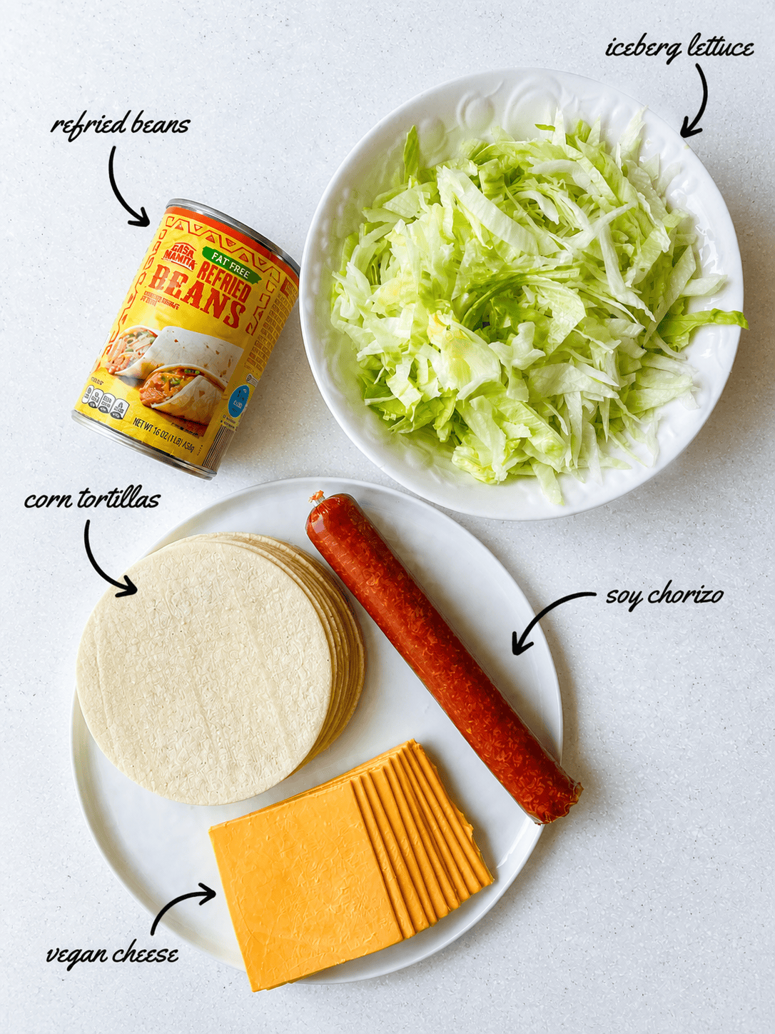Ingredients for vegan Jack in the Box tacos copycat recipe, including refried beans, iceberg lettuce, soy chorizo, corn tortillas, and vegan cheese, labeled in an overhead view.