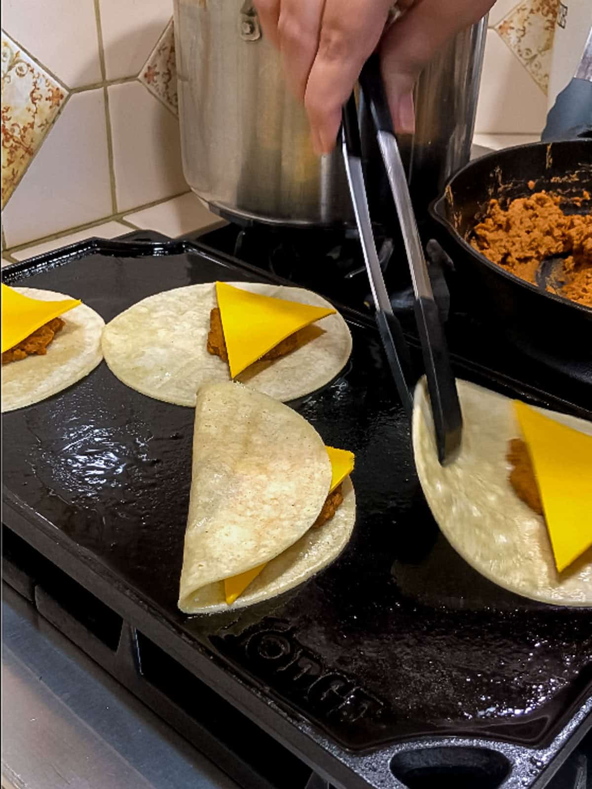 Folding corn tortillas over vegan taco filling and cheese using tongs on a hot griddle.